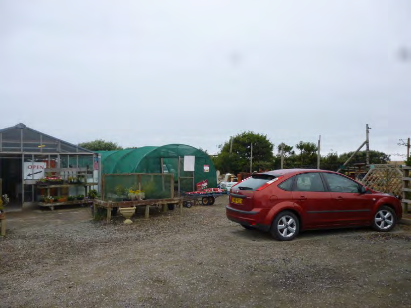 A photograph of a rural gravel yard featuring a glasshouse, a plastic tunnel greenhouse, and a parked red car.