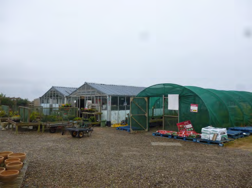 A photograph showing an existing agricultural plot featuring glass greenhouses and a large green polytunnel on a gravel surface.