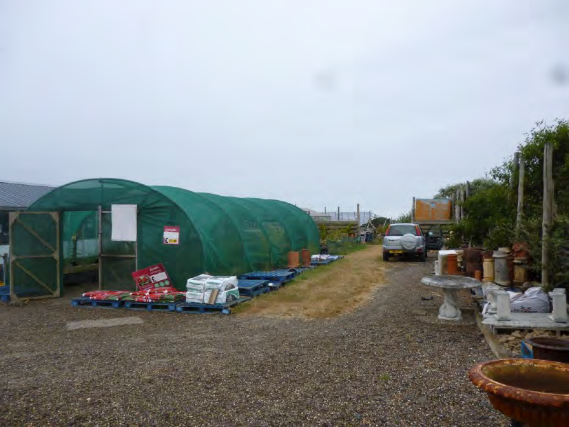 A photograph showing a large green polytunnel greenhouse on a gravel plot, surrounded by gardening supplies, pots, and a vehicle.