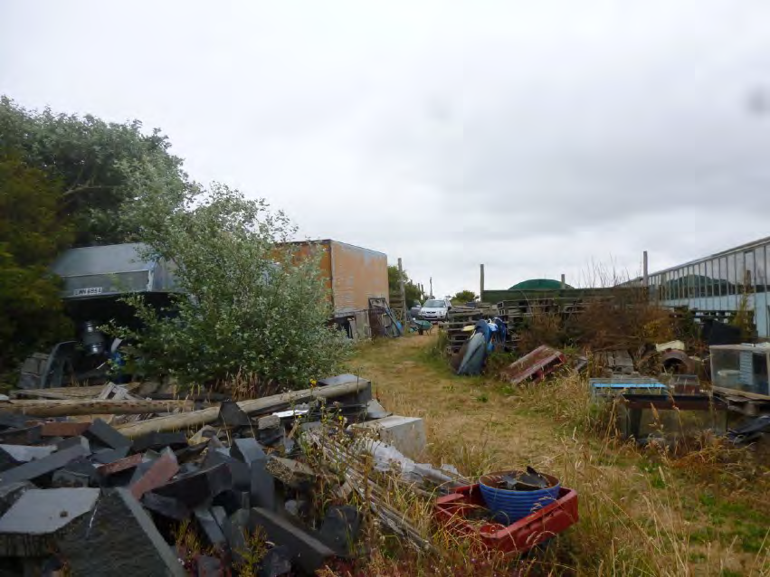 A photograph of a cluttered, overgrown plot of land featuring piles of slate debris, old wooden structures, and scattered items in a rural setting.