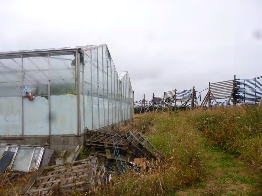 A photograph showing a large, weathered greenhouse structure with a metal frame and glass panels. The foreground contains debris and wooden pallets, while a grassy slope with a wooden windbreak fence is visible to the...