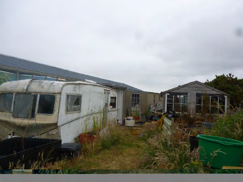 A photograph showing a dilapidated caravan and overgrown garden area with adjacent outbuildings and greenhouse structures.