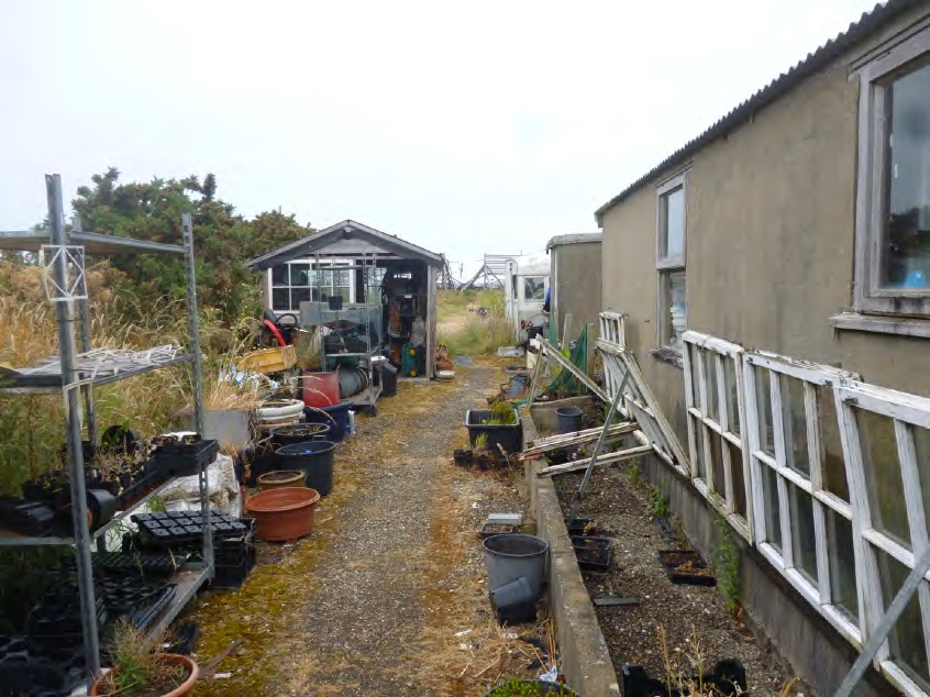 A photograph showing a cluttered garden or yard area next to a single-story building, featuring gardening supplies, pots, and a small shed in the background.