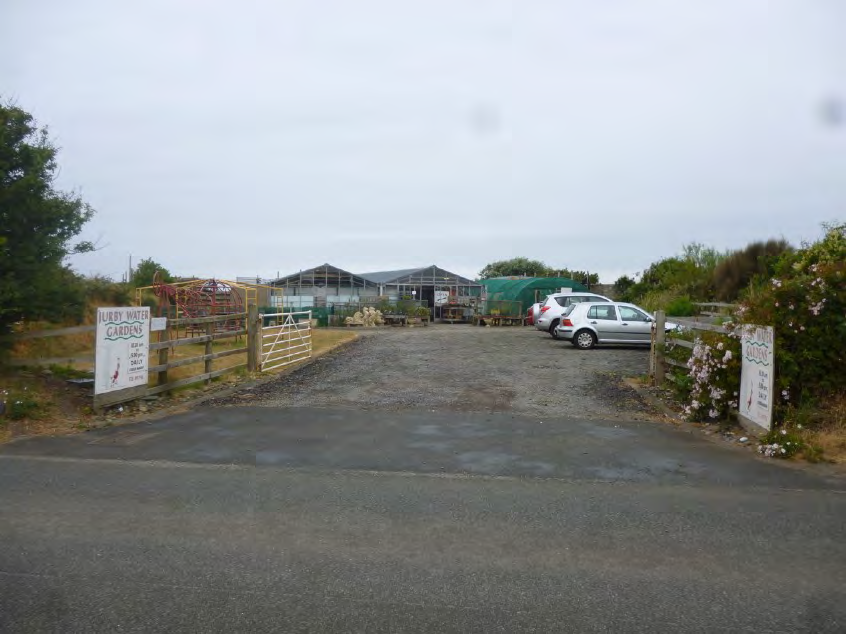A photograph showing the entrance to a rural property with a gravel driveway, wooden gates, and greenhouse structures in the background.