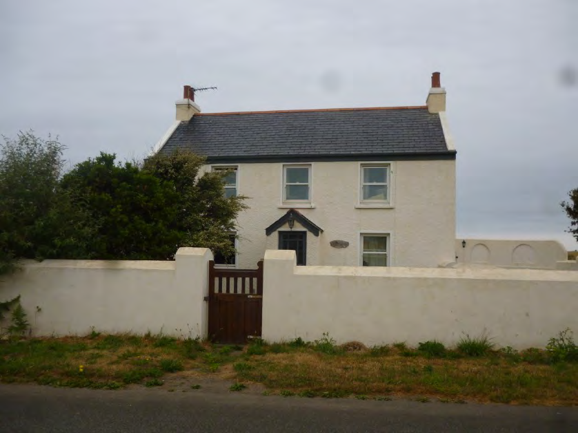 A photograph showing a white, two-story detached house with a slate roof, located behind a white boundary wall and wooden gate.
