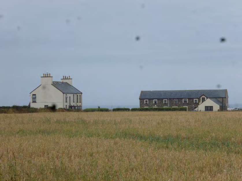 A photograph showing a rural landscape with a grassy field in the foreground and two buildings, a white house and a long stone structure, in the background.
