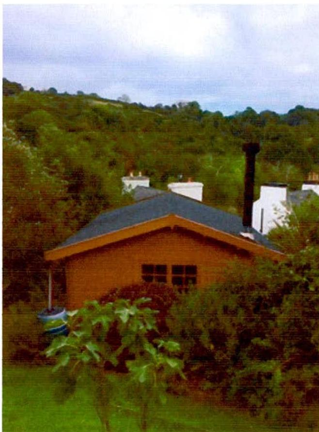 A photograph showing a wooden summerhouse or small cabin with a slate roof and chimney, situated in a green, hilly environment.