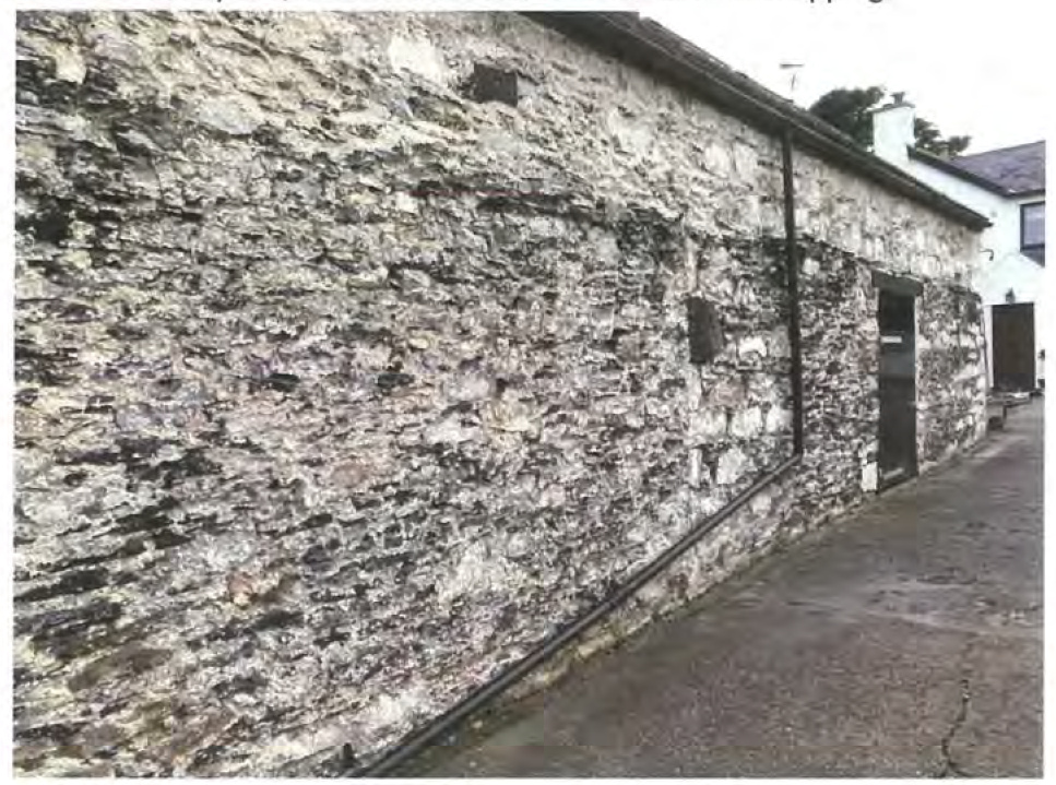 A photograph showing the rough stone exterior wall of a long outbuilding, likely a barn, with a paved driveway alongside.