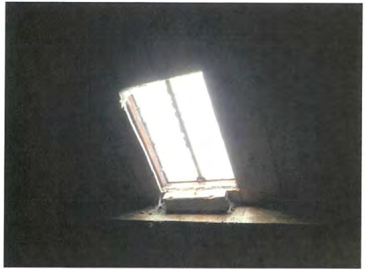 A grainy photograph showing the dark interior of a loft or attic space with a bright skylight window set into the sloped ceiling.