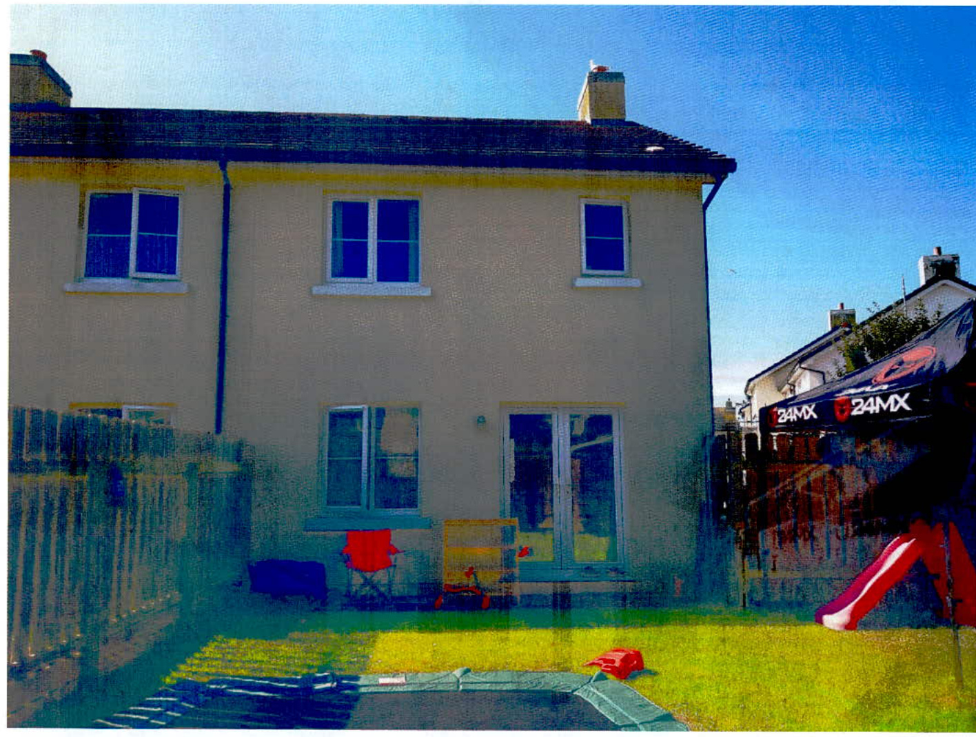 A photograph showing the rear elevation of a two-story residential house with a lawn, garden furniture, and a wooden fence.