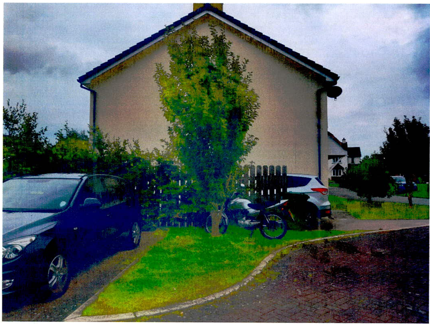 A photograph showing the side elevation of a two-story detached house with a driveway, parked cars, and a tree in the front garden.
