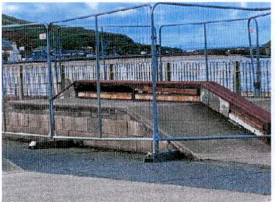 A photograph showing a temporary metal fence enclosing a concrete ramp or platform structure near a body of water.