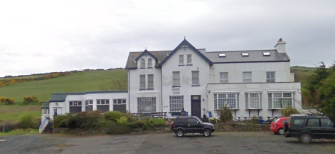 A photograph showing a large white multi-story building, identified as a former hotel, situated in a rural setting with a parking area in the foreground.