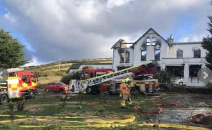 A photograph showing a large white building, likely the former hotel, engulfed in smoke and fire with multiple fire engines and firefighters attending the scene.