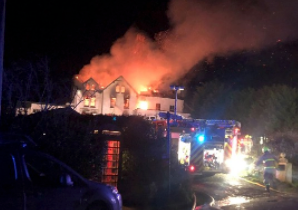 A night-time photograph showing a large building, likely the former hotel, engulfed in flames with fire trucks and emergency personnel on the scene.