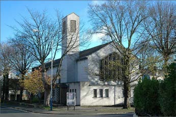 An exterior photograph of a white building with a tall tower, surrounded by bare trees and a signpost, likely the existing site.