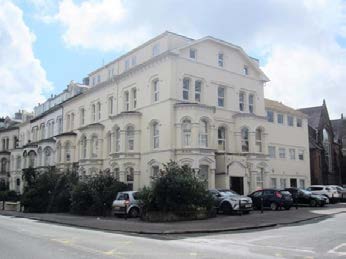 A photograph showing the exterior of a large, white, multi-story building with classical architectural features, likely the proposed multipurpose hall, with cars parked in the foreground.
