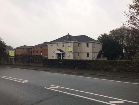A street-level photograph showing a large, light-colored building with a dark roof situated behind a stone wall along a road.