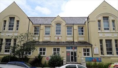 A photograph showing the exterior of a large, cream-colored building with a central entrance canopy and gabled roof sections, with cars parked in the foreground.