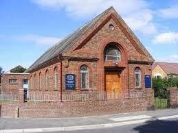 A street-level photograph of a red brick building with a pitched roof and arched windows, appearing to be a community hall or chapel.