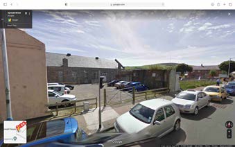 A street-level photograph showing a parking area with several cars and a large building with a pitched roof in the background, likely the site of the proposed development.