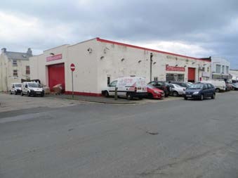 A photograph showing the exterior of a large, white single-story commercial building with red roller shutter doors and red roof trim. Several vehicles are parked in front on a paved surface.
