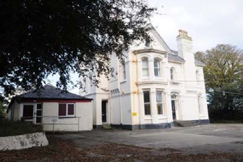 A photograph showing a white two-story building with a single-story extension on the left and a paved parking area in the foreground.