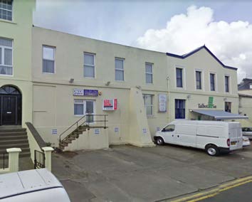 A street-level photograph showing a cream-colored building complex with a paved forecourt where white vans are parked.