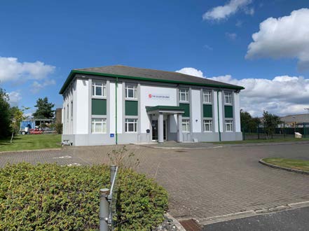 A photograph showing a white community building with green window frames and an entrance canopy, situated next to a paved parking area under a blue sky.