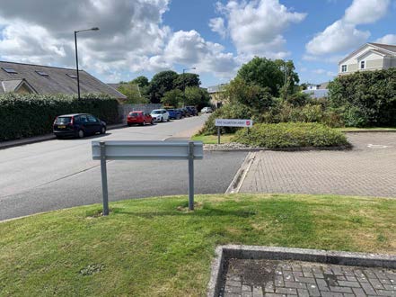 Ground-level photograph showing a paved parking area, a road with parked cars, and surrounding vegetation and buildings.
