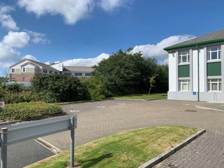 A photograph showing a paved parking area in front of a white building with green trim and another building in the background under a blue sky.