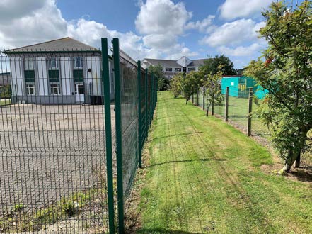 A photograph showing a large white building with a grey roof situated behind a green metal fence and a gravel forecourt area.