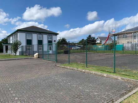 A photograph showing a white two-story building with a large paved car park in the foreground and a fenced sports area with a slide in the background.