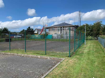 A photograph showing a building under construction with scaffolding, likely the proposed multipurpose hall. The foreground features a paved parking area and a fenced sports court.
