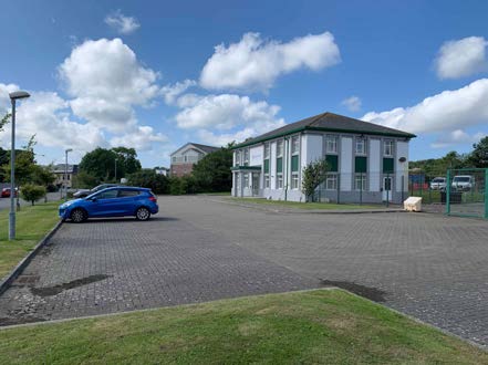 A photograph showing a white two-story building with green shutters and a large paved parking area in the foreground.