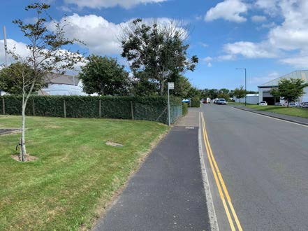 A street-level photograph showing a paved road with double yellow lines, a sidewalk, and a grassy verge with a fence and trees. Industrial or commercial buildings are visible in the background under a blue sky.