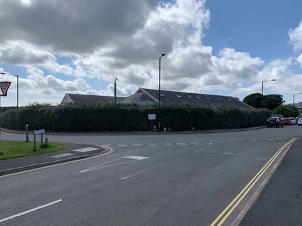 A street-level photograph showing a large building with a pitched roof situated behind a tall green hedge along a roadside.