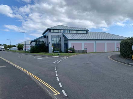 A photograph showing a large, single-story building with a grey pitched roof and pink side panels, situated next to a paved road with double yellow lines.