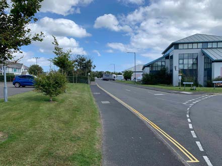 A street-level photograph showing a modern building with a large glass facade on the right side of a road, likely the proposed multipurpose hall, with a blue car parked on the left and a bus in the distance.