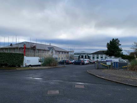 A photograph of a large single-story building undergoing renovation with scaffolding on the left, a white van, and a paved car park area in the foreground.