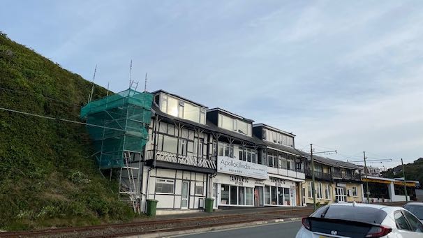 A street-level photograph showing a row of terraced buildings with scaffolding and green netting covering the upper section of the leftmost building, indicating construction work near a railway line.