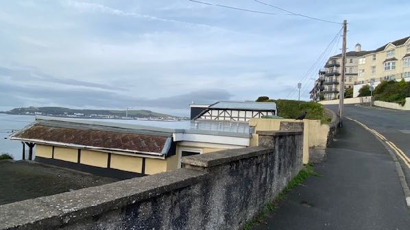 A street-level photograph showing a single-story building with a flat glass-roofed extension near the coast, with a larger residential building on a hill in the background.