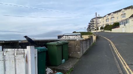 A street-level photograph showing a coastal road curving uphill with large residential buildings in the background and the sea visible to the left.