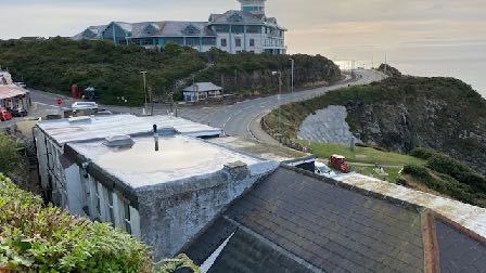 A high-angle photograph showing a flat roof in the foreground, a coastal road curving along a cliff, and a large building overlooking the sea in the distance.
