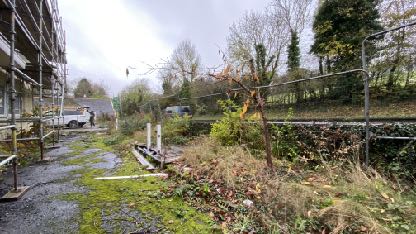 A photograph showing a building covered in scaffolding on the left side, indicating ongoing construction or renovation work. The foreground features a muddy area with wooden planks and a wire fence separating the site...