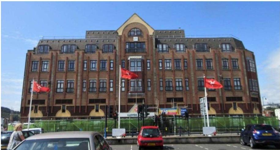 A photograph of a large, multi-story brick commercial building with numerous windows and Isle of Man flags flying in the foreground.
