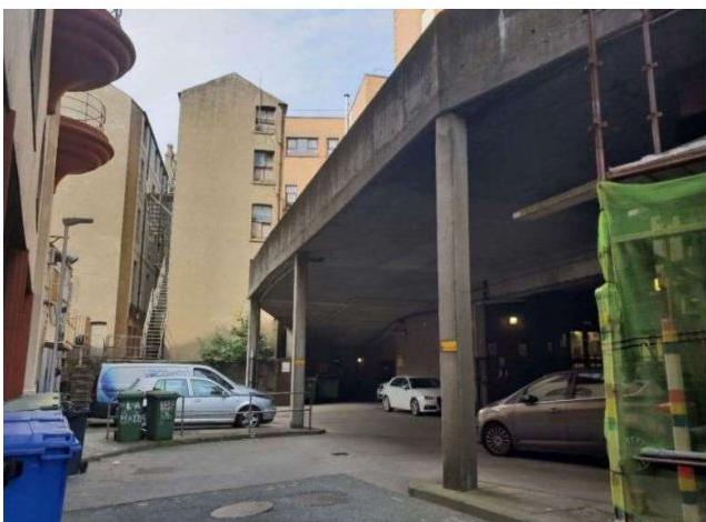 A street-level photograph showing a large concrete structure, likely a multi-story car park, supported by thick pillars. Several cars are parked underneath, and urban buildings with balconies are visible in the backgr...