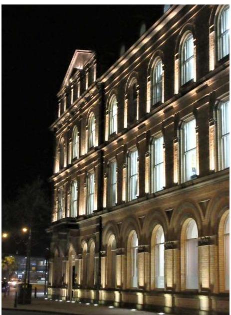 A night-time photograph of a large, multi-story brick building featuring arched windows and illuminated facade uplighting.