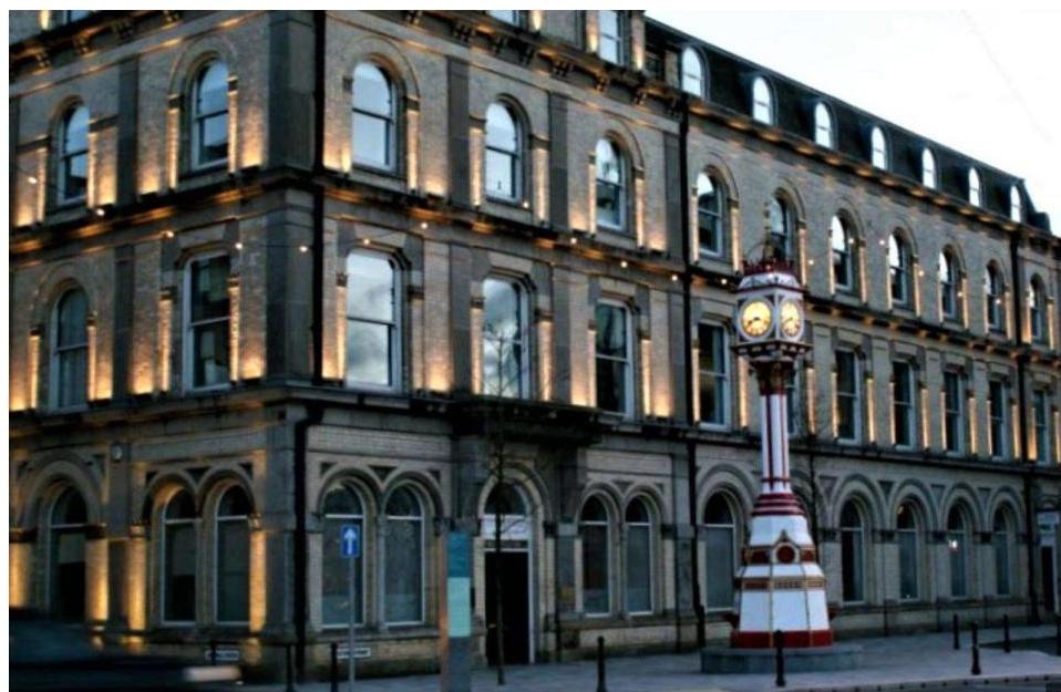 A photograph showing the exterior facade of a large, multi-story stone building illuminated by uplighting. A distinctive clock tower stands in the foreground on the pavement.
