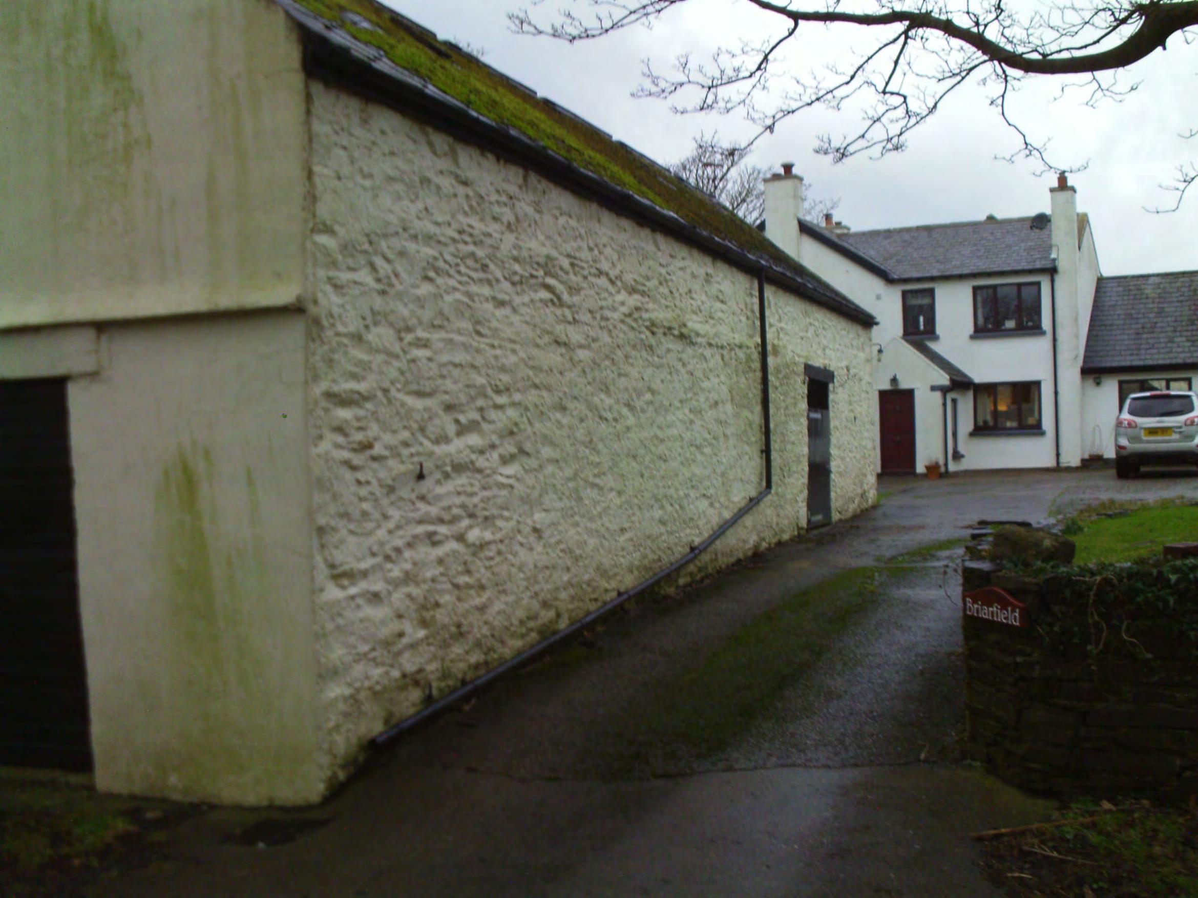 Exterior photograph showing a long white-washed stone building, likely the existing barn, alongside a driveway leading to a residential house.